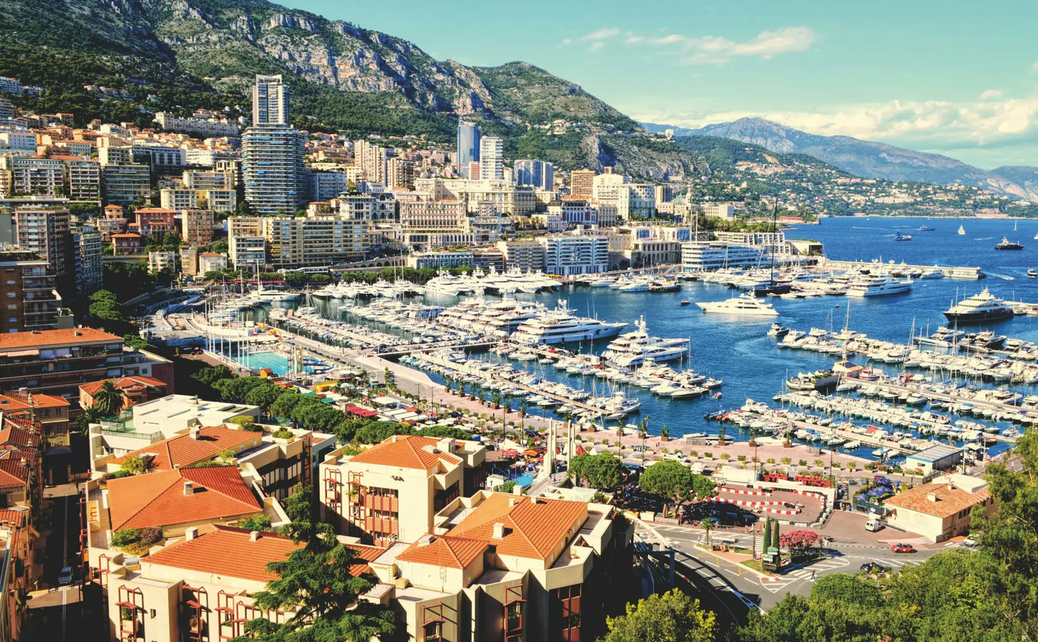 A bustling harbor in monaco, filled with numerous white and silver yachts docked among terracotta-roofed buildings nestled against green mountains.