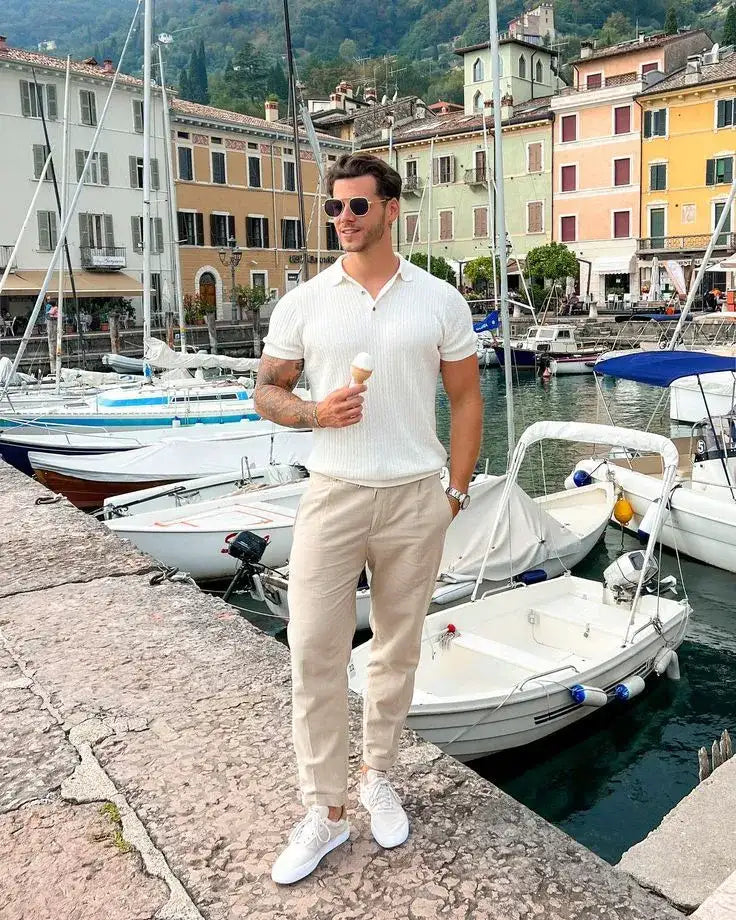A man wearing a white ribbed polo shirt, beige chinos, and white sneakers stands by the water with boats in the background.